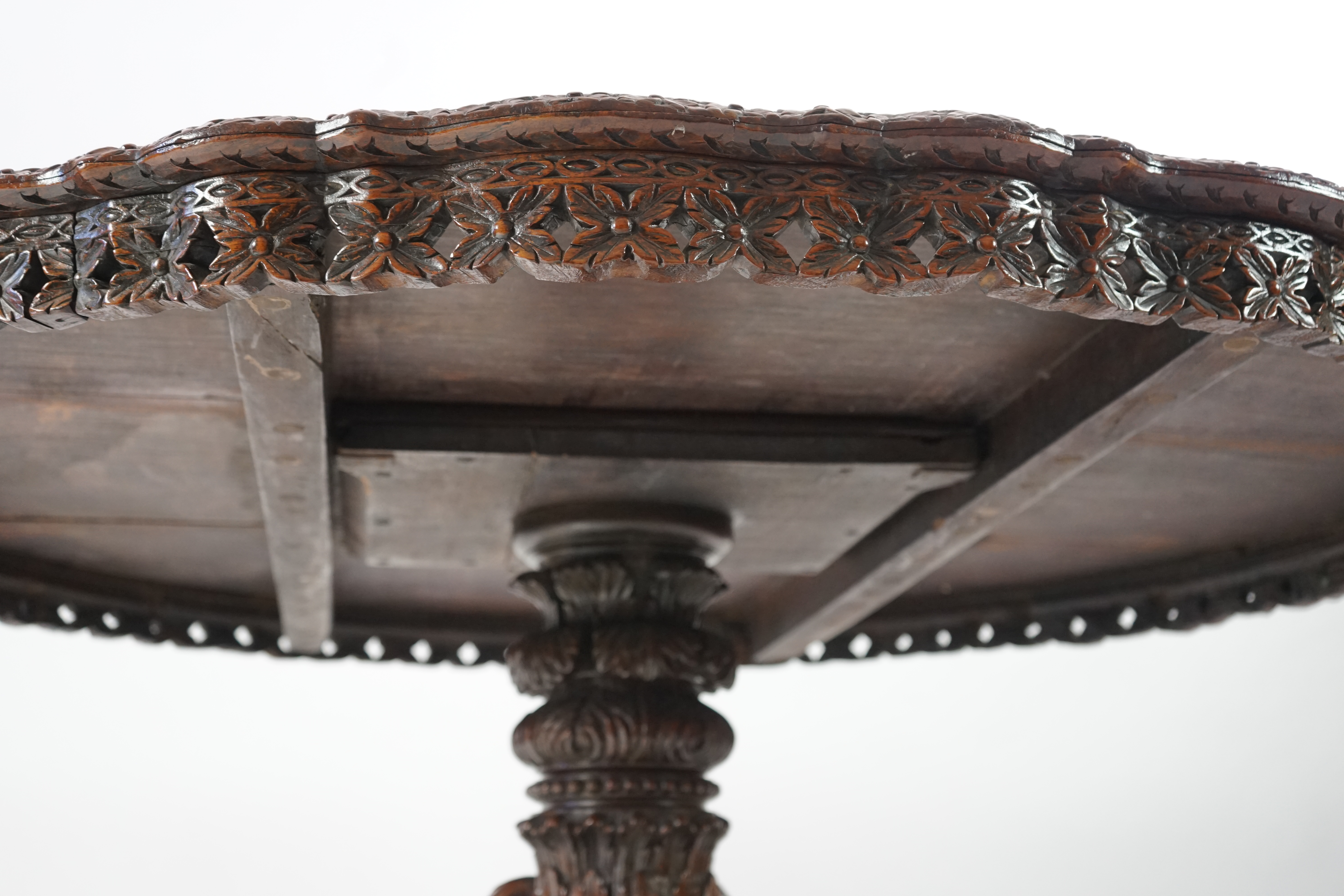 A 19th century Anglo-Indian hardwood foliate carved tilt top centre table, with serpentine circular top, stylised floral carved rim and frieze, on heavily foliate and fruit carved pillar with pierced triangular platform